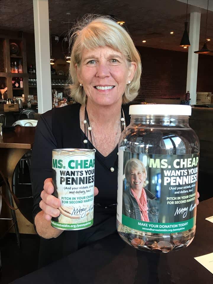 A smiling woman with short blonde hair holds a donation can and a large jar labeled Ms. Cheap Wants Your Pennies, encouraging people to donate spare change for a fundraiser.