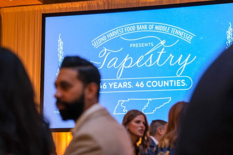 People gather in front of a large blue screen that reads, “Second Harvest Food Bank of Middle Tennessee presents Tapestry: 46 Years. 46 Counties,” with outlines of Tennessee at the bottom.