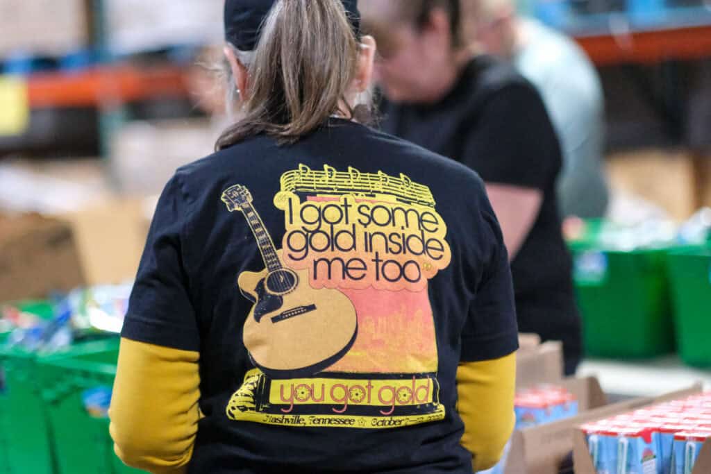 A person wearing a black t-shirt with a guitar graphic and the words “I got some gold inside me too. You got gold” stands in front of a table with green bins and food items, working with others in a warehouse.