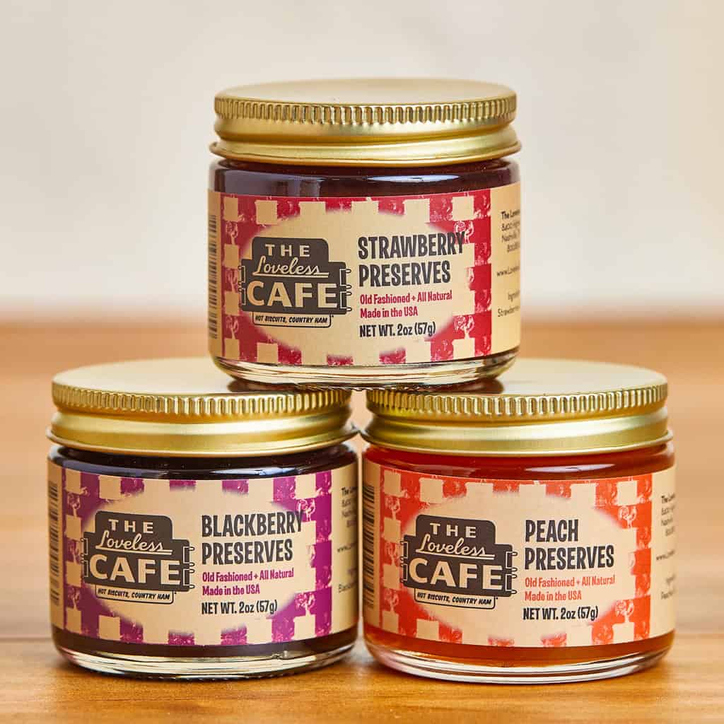 Three small glass jars of fruit preserves are stacked on a wooden surface. The labels read Strawberry Preserves, Blackberry Preserves, and Peach Preserves from The Loveless Café.