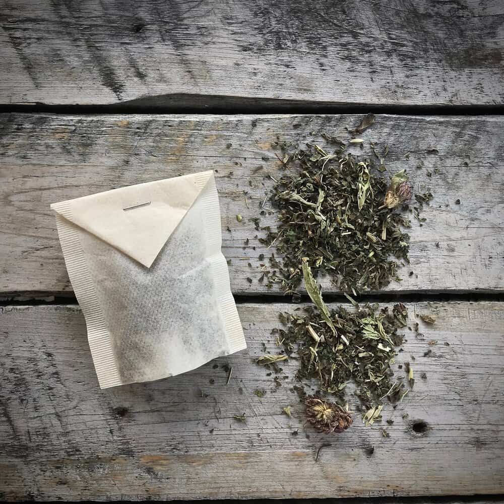 A tea bag and a pile of loose dried herbs are placed on a rustic wooden surface. The tea bag is sealed, while the loose herbs show a mix of green leaves and small flower pieces.