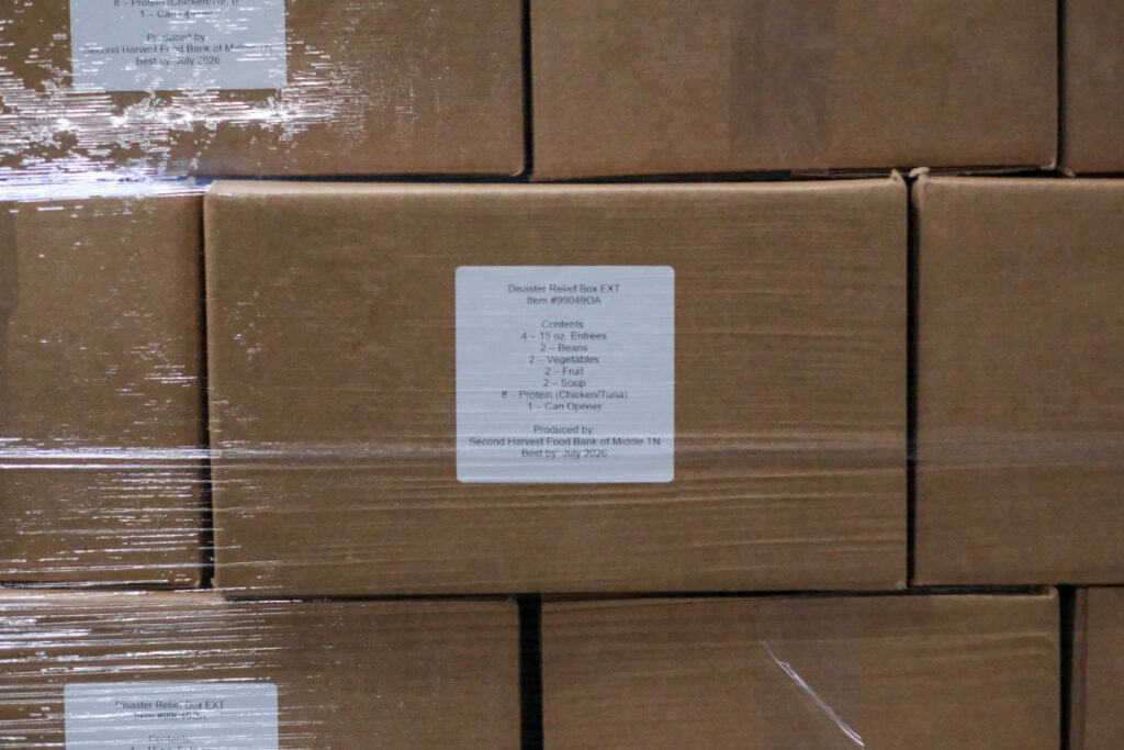 A close-up of stacked cardboard boxes wrapped in plastic. One box, labeled Second Harvest disaster relief, lists contents for a Disaster Relief Box, including entrees, fruit, snacks, and soup.