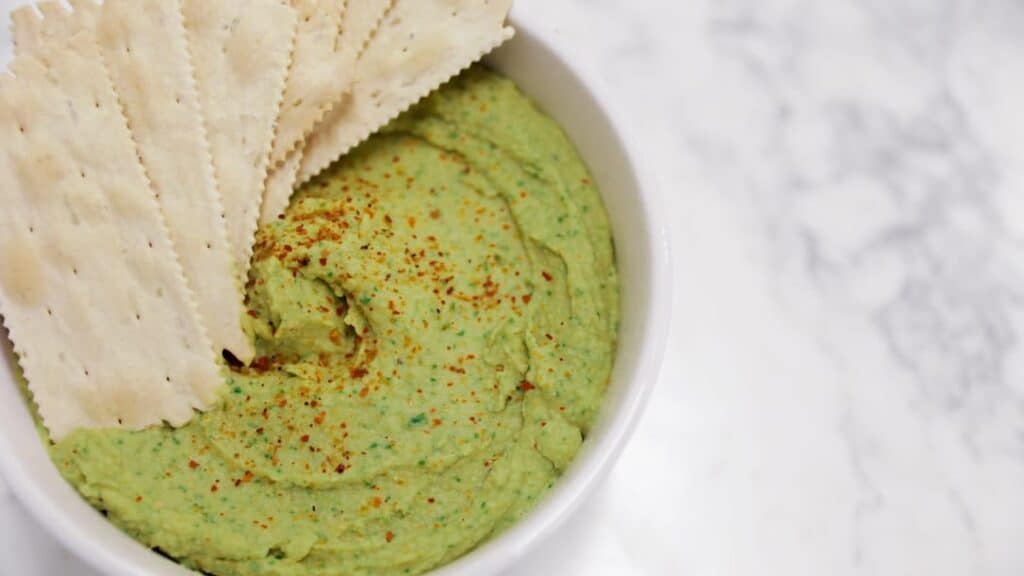 A white bowl filled with green hummus sprinkled with spices, served with several rectangular crackers on top, set on a white marble surface.
