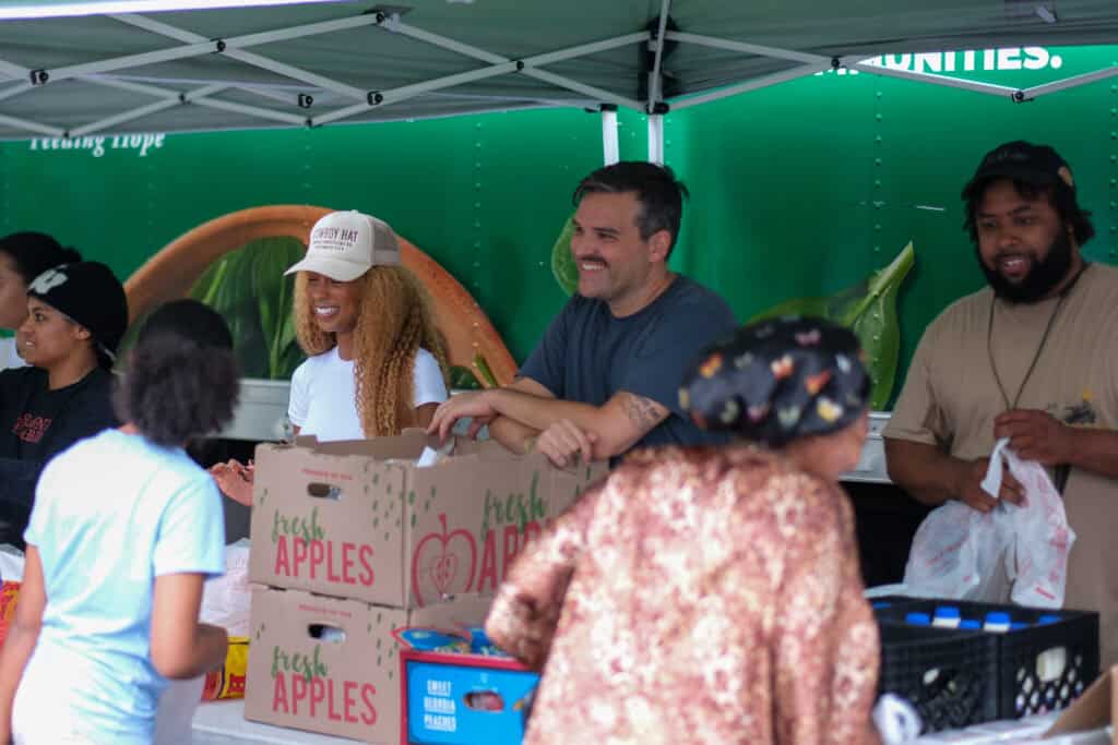 Volunteers smile and distribute boxes of fresh apples to people at an outdoor community food event, standing under a canopy with a green background.