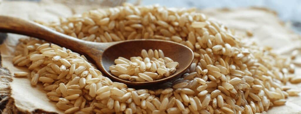A wooden spoon rests on a pile of uncooked brown rice grains scattered on a piece of parchment paper.