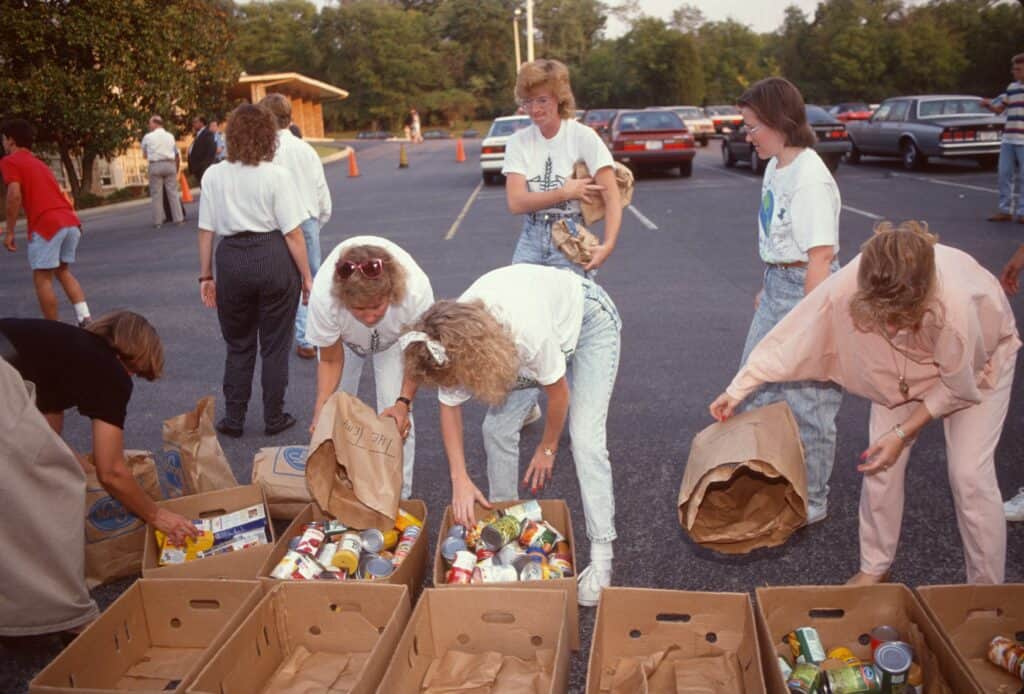A group of people outdoors sorts canned goods into paper bags and boxes in a parking lot, likely organizing donations or at a food drive. Cars are parked in the background.