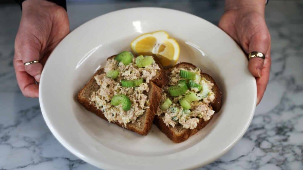 A plate with two slices of bread topped with tuna salad and celery pieces, garnished with two lemon slices. Two hands with rings are holding the plate over a marble surface.