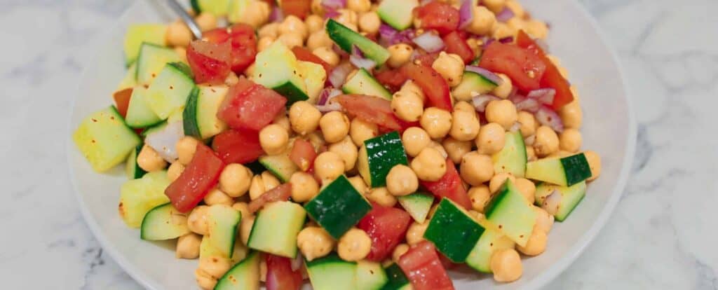 A white plate filled with a colorful salad made of chickpeas, cucumber, tomato, and chopped red onion, all tossed together and lightly seasoned.