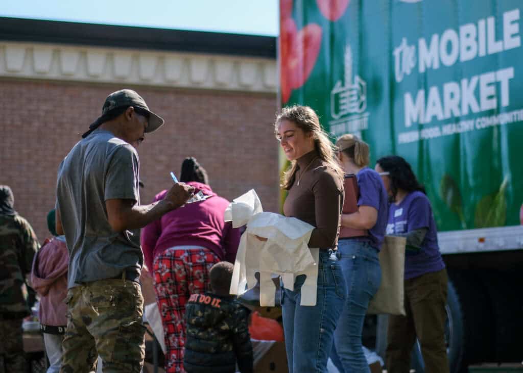 A group of people gather outside near a large truck labeled The Mobile Market. A woman smiles while holding a paper bag, and others stand nearby, some collecting or distributing items.