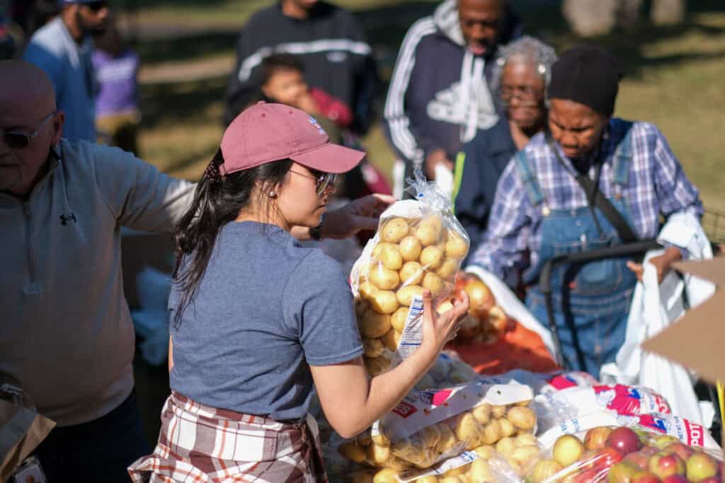 A woman in a pink cap hands out bags of potatoes to people gathered at an outdoor food distribution event. Several people wait in line, holding bags and carts, on a sunny day.