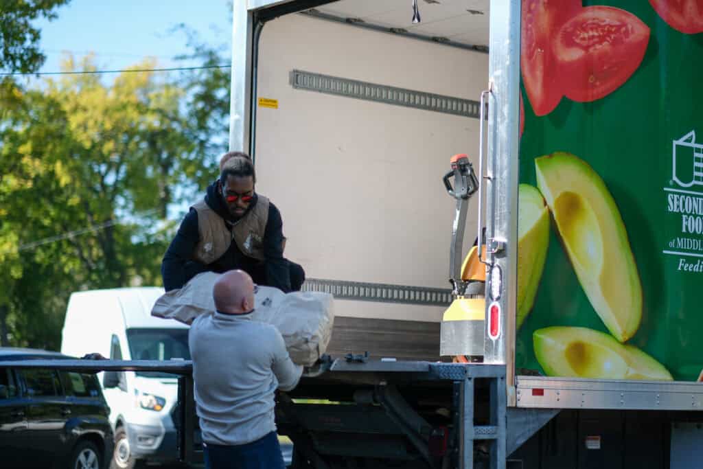 Two people unload a large bag from a truck with images of tomatoes and avocados on its side. One person stands inside the truck handing the bag to another person waiting outside. Trees and parked vehicles are in the background.