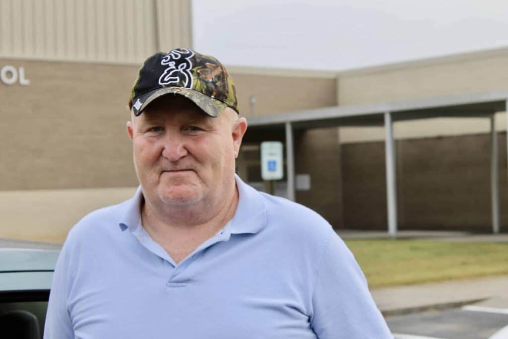 An older man in a blue polo shirt and camouflage hat stands outside near a beige brick building with a covered walkway and a sign in the background.