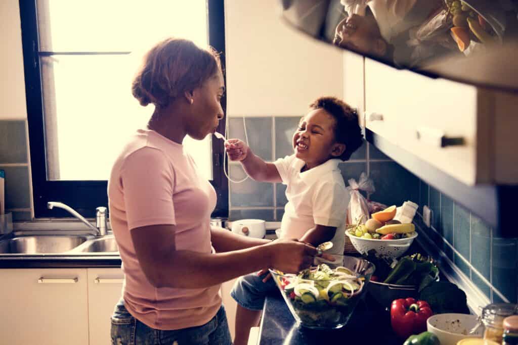 A woman and a young child are in a bright kitchen. The woman is preparing a salad while the smiling child, sitting on the counter, offers her a spoonful of food. Fresh vegetables and fruit are visible nearby.
