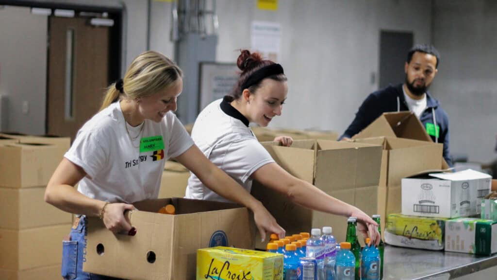 Three people pack boxes with drinks and supplies at a table in a warehouse. Two women smile while filling boxes, and a man stands nearby. Bottles of water and sports drinks are visible on the table.