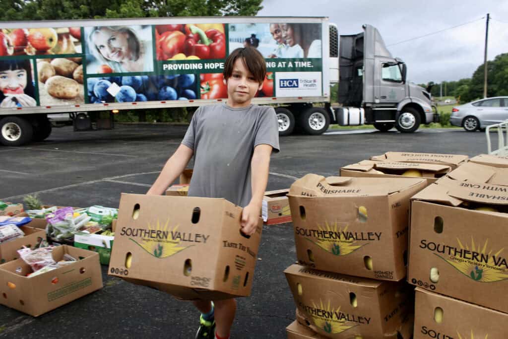 A young boy carries a large box labeled Southern Valley in a parking lot, surrounded by other boxes of produce. In the background, a truck with images of fruits, vegetables, and smiling faces is parked.