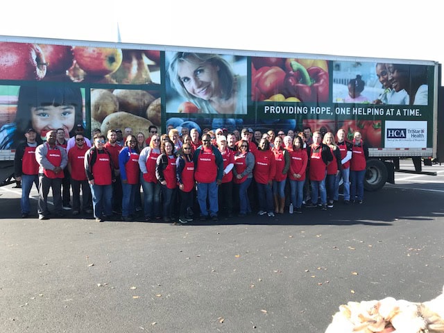 A large group of people wearing matching red vests pose in front of a truck with images of produce and smiling faces, showing their commitment to Fighting Hunger this Holiday with Nissan. The text on the truck reads, “Providing hope, one helping at a time.”.