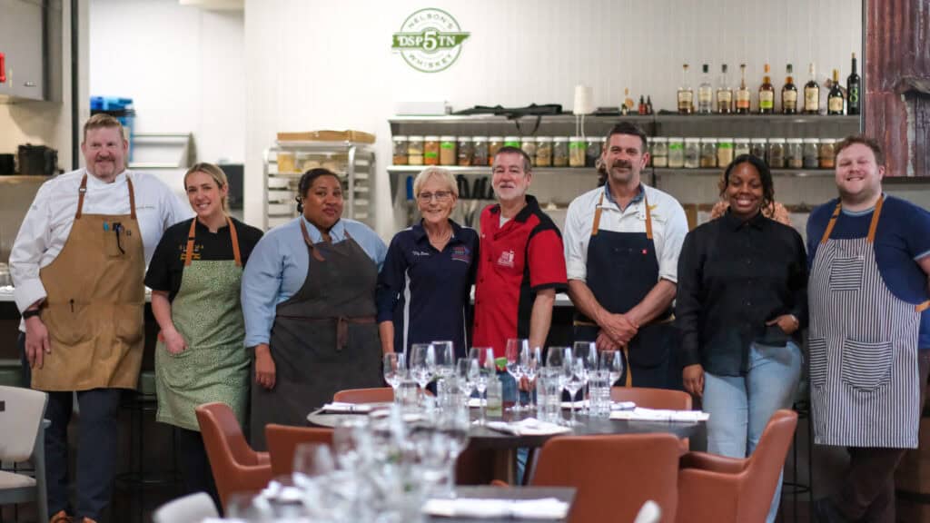 Eight people, including chefs in aprons and restaurant staff, stand and smile together in a restaurant kitchen with wine glasses and dining tables in the foreground. Shelves and bottles are visible in the background.