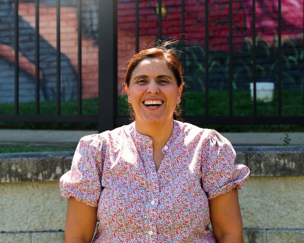 Claudie, who knows how poverty can impact a person's life, smiles in a floral blouse. She sits outside by a colorful mural in front of a Second Harvest Partner Agency, Catholic Charities.
