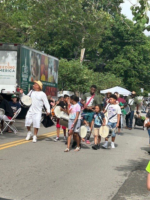 A group of children and an adult walk down a street playing drums during an outdoor event, with trees, tents, and a Feeding America truck in the background. Spectators sit and watch along the sides of the street.