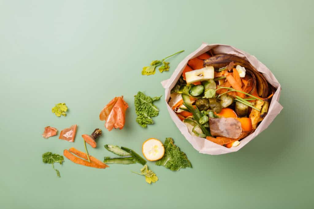 A paper bag filled with assorted vegetable scraps and food waste sits on a green surface, showing simple ways to help on Earth Day by composting leftovers, with some peelings and scraps scattered around it.