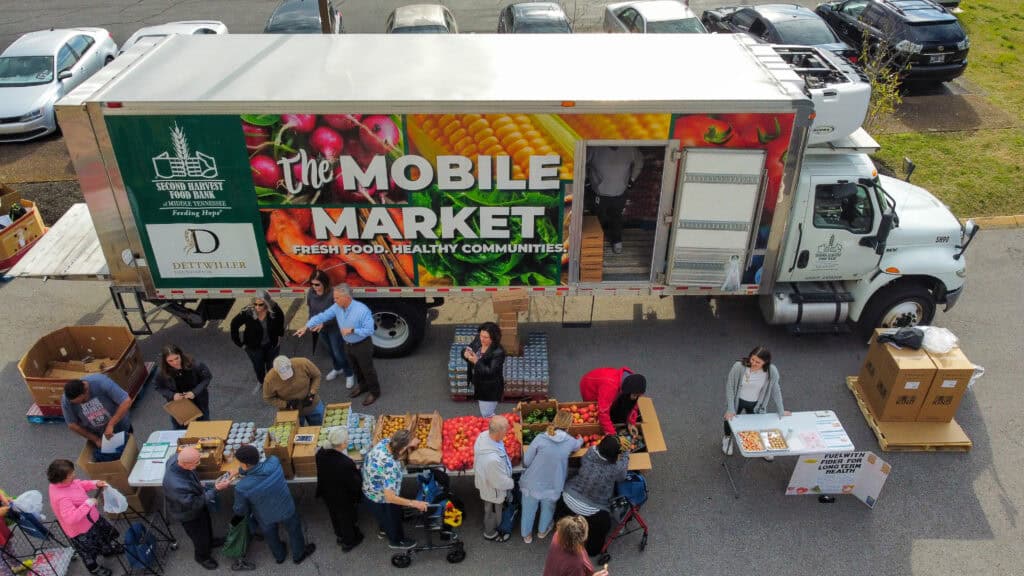Aerial view of a colorful mobile market truck with Fresh Food, Healthy Communities written on its side. People gather at tables in front of the truck, selecting fresh produce and interacting with vendors in a parking lot. Shows how Second Harvest is Building Sustainable Food Systems.