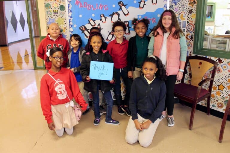 A group of eight smiling children pose indoors in front of a decorated penguin-themed bulletin board. One child holds a blue sign that says “Thank you.” They are casually dressed and appear happy.