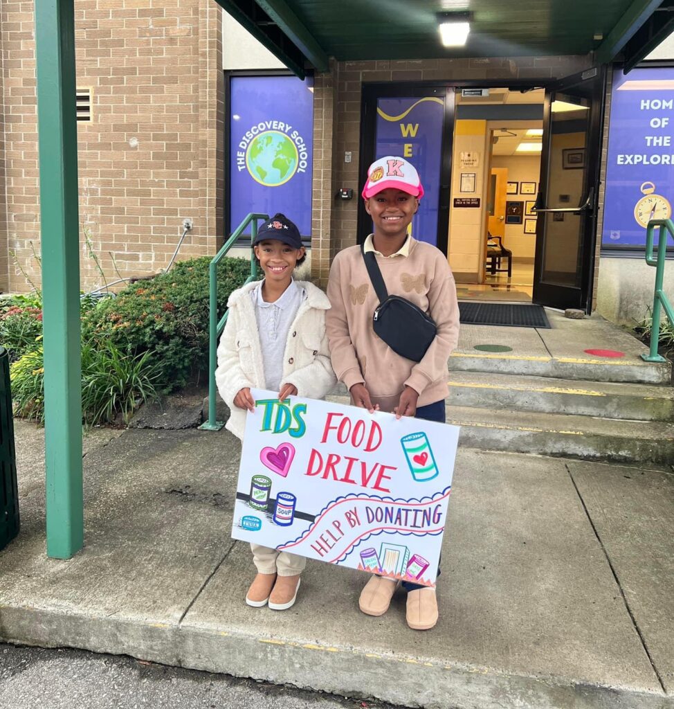 Two children stand outside a building entrance, smiling and holding a colorful handmade sign that reads “TDS Food Drive—Help by Donating,” with illustrations of cans and jars. Both are wearing jackets and baseball caps.