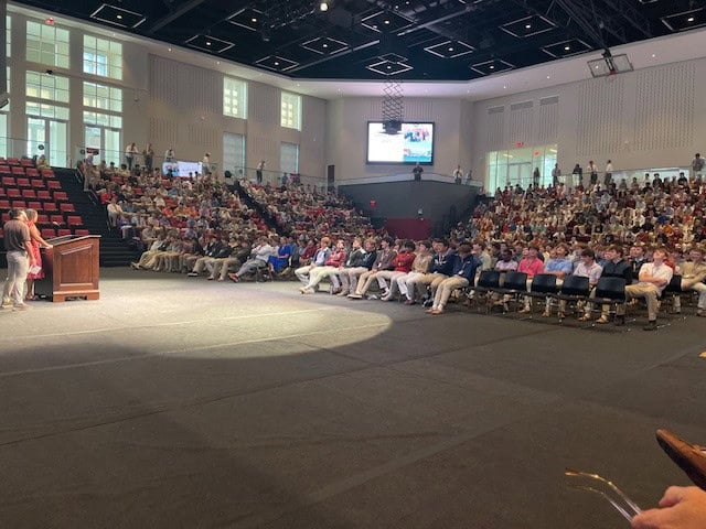 A large audience sits in a modern auditorium with tiered seating, listening to a speaker standing at a podium on stage; the room is well-lit with many people attentively watching.