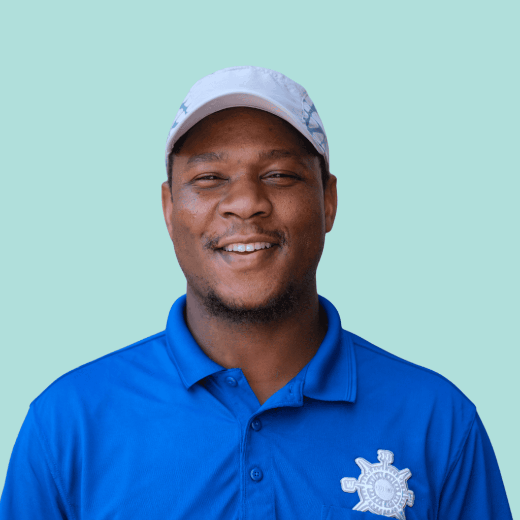 Smiling man wearing a blue polo shirt with an embroidered logo and a light-colored cap, standing in front of a solid light blue background.