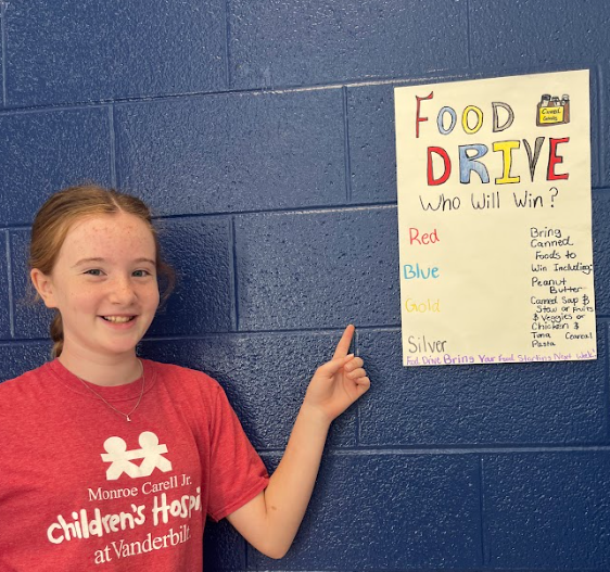 A girl in a red Monroe Carell Jr. Children’s Hospital at Vanderbilt t-shirt smiles and points to a handmade “Food Drive” poster taped to a blue wall. The poster lists team colors and donation instructions.