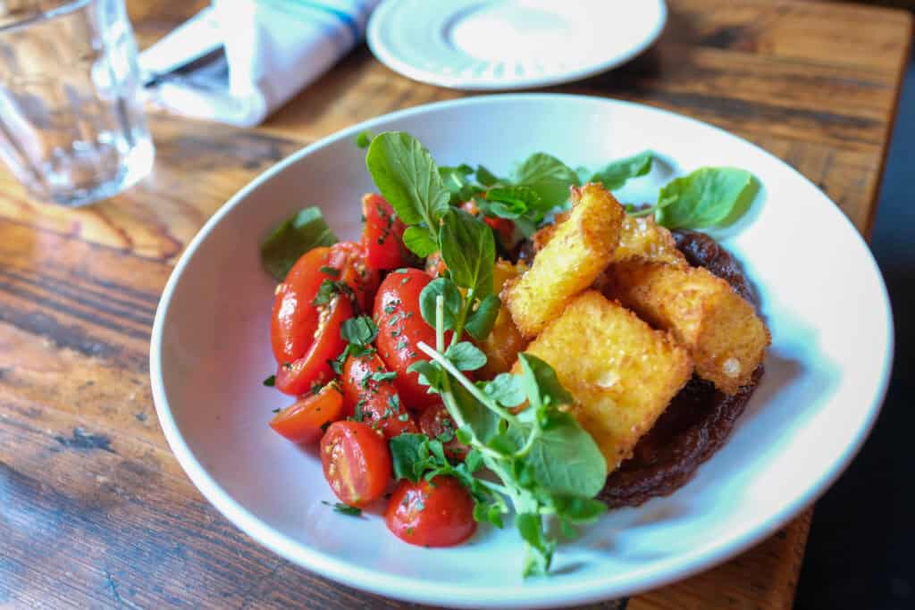 A white bowl with crispy fried polenta sticks, halved cherry tomatoes, fresh greens, and herbs sits on a rustic wooden table next to a glass and a plate with a napkin.