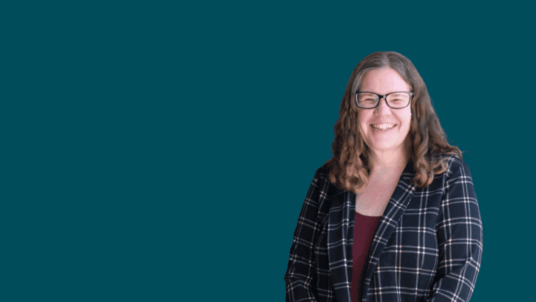 A smiling woman with long wavy hair, wearing glasses, a dark checkered blazer, and a maroon top, stands against a plain teal background.