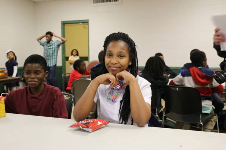 A girl sits at a table smiling, holding a juice pouch, with a bag of chips in front of her. Other children are sitting and standing in the background in a classroom or community room setting.