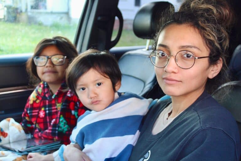 A woman with glasses sits in a car holding a young child wrapped in a striped blanket, with another child in glasses and a red plaid jacket sitting beside them. All three look toward the camera.