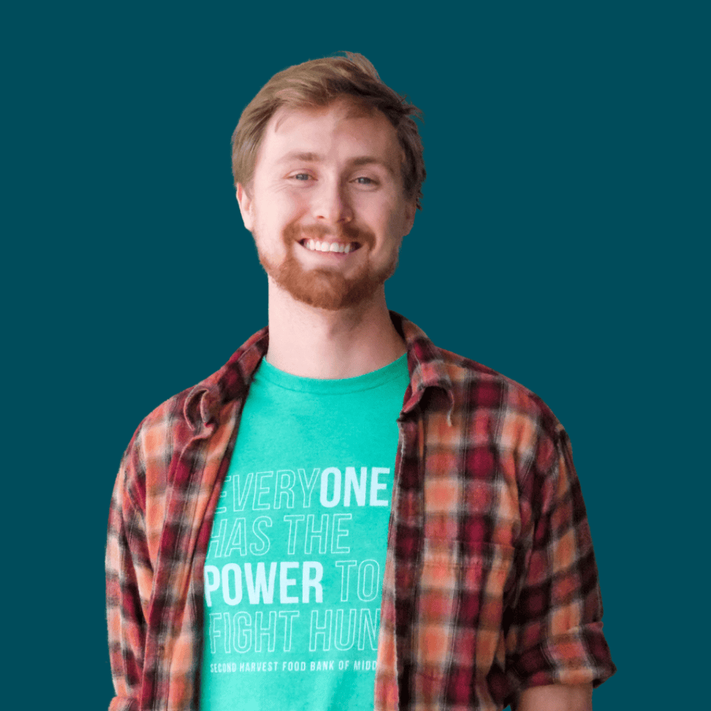 A smiling young man with light brown hair and a beard wearing a green T-shirt and an open red flannel shirt, standing in front of a solid teal background.