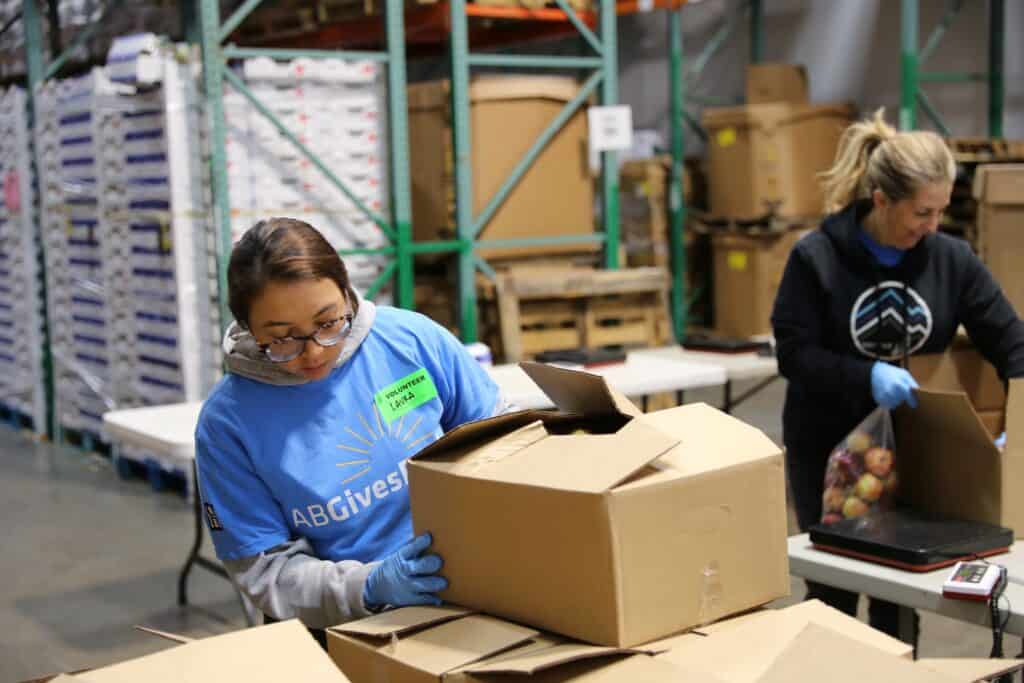 AllianceBernstein Invests in a Brighter Future at Second Harvest. Two volunteers wearing gloves pack food items into cardboard boxes at a warehouse. One person in a blue shirt handles a box, while another in a black hoodie fills a box with apples. Shelves and supplies are visible in the background.