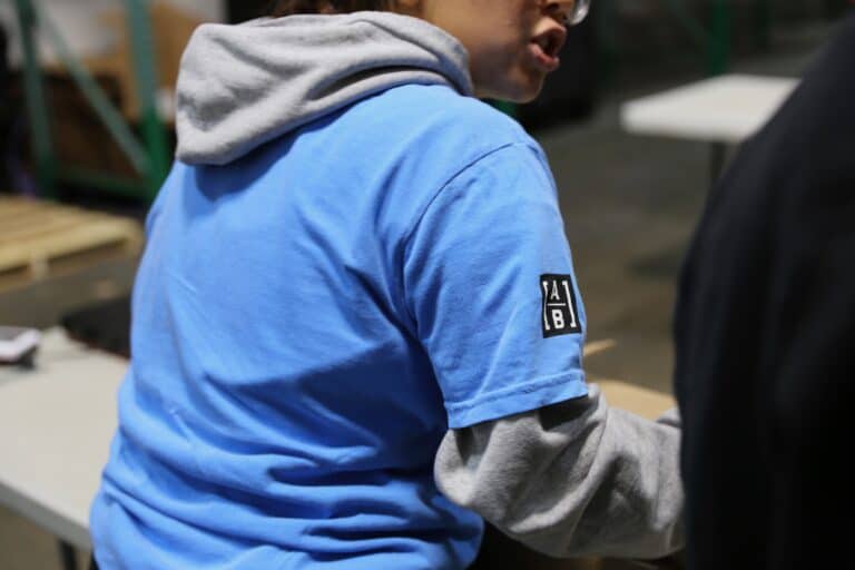 A person wearing a light blue T-shirt over a gray hoodie sits at a table in an indoor setting. The T-shirt has a black and white patch on the sleeve, and the persons face is partly out of view.