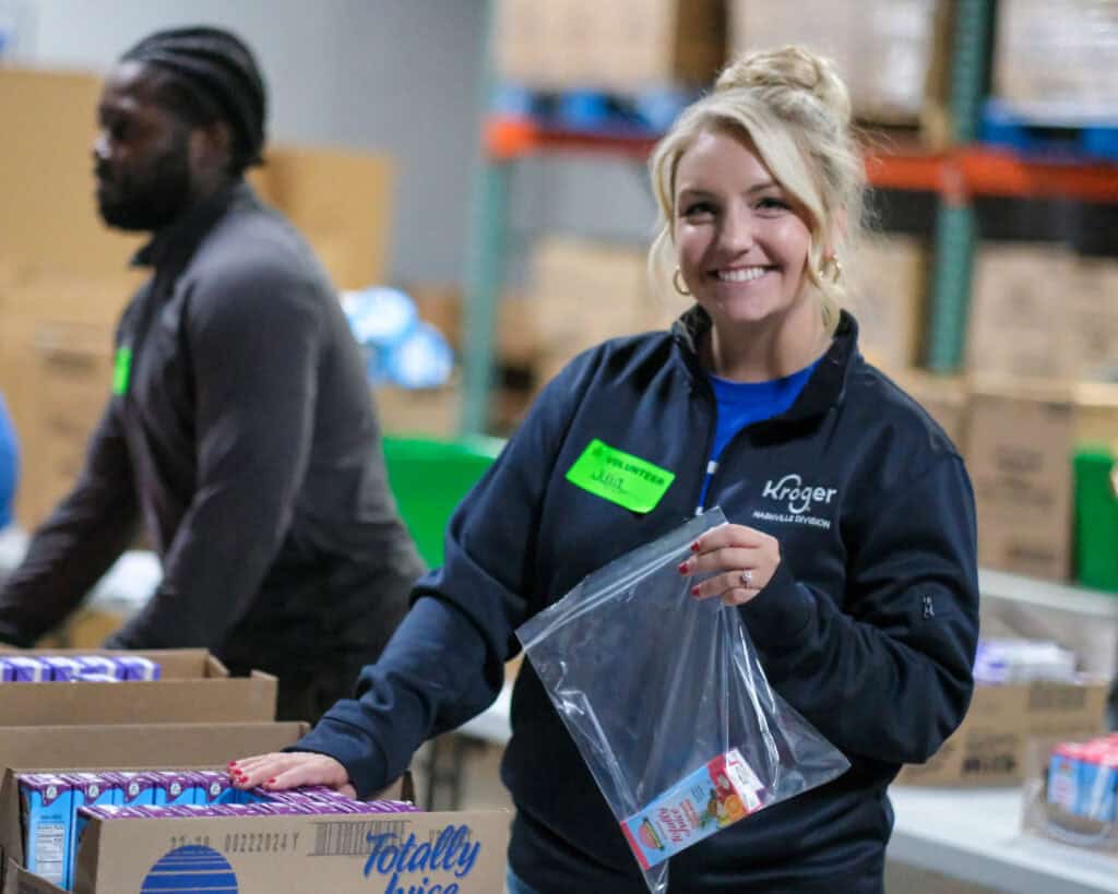 A woman smiles while packing food into a plastic bag at a warehouse, proudly wearing a Kroger jacket and name tag. In the background, another person works—demonstrating how Kroger multiplies their impact with the Power of ONE.