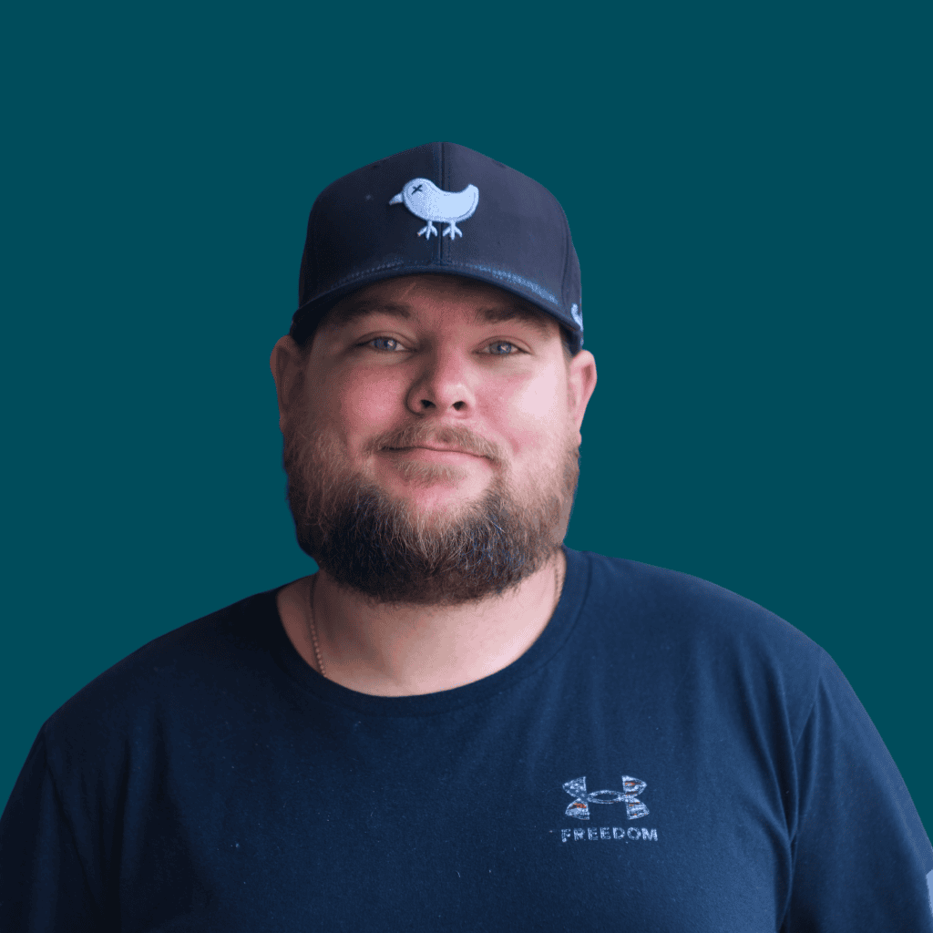 A man with a beard wearing a black baseball cap featuring a bird logo and a black T-shirt stands in front of a solid teal background, smiling slightly at the camera.