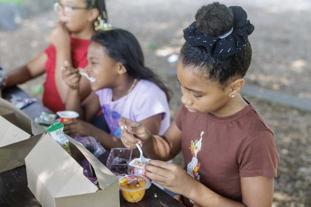 Three school-aged children sit at an outdoor table eating yogurt, fruit cups, and sandwiches from lunch boxes. The image illustrates how to pack healthy lunches for kids using balanced, affordable foods.