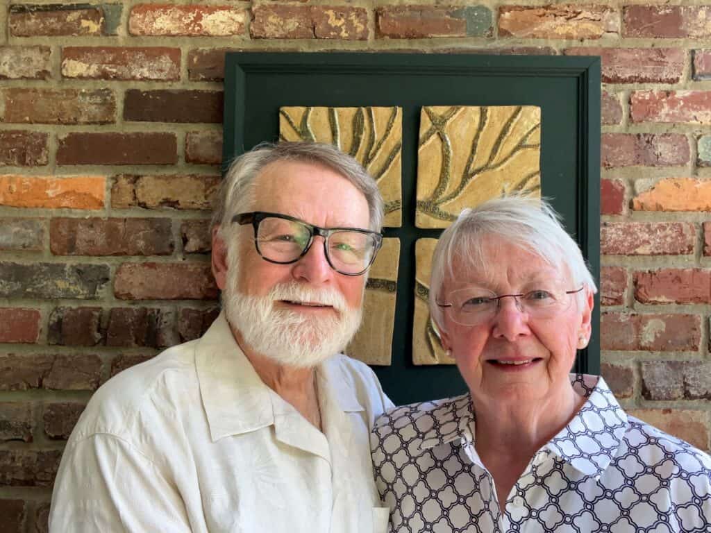 Considering Planned Giving with Life Insurance? An older man and woman stand close together, smiling at the camera in front of a brick wall and framed artwork. The man has white hair, a beard, and glasses; the woman has short white hair and glasses.