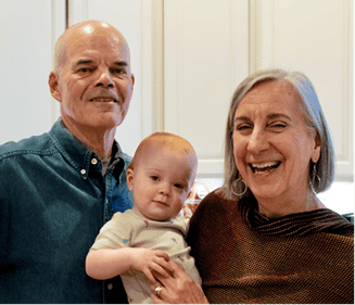 Kren, featured in Second Harvest’s blog “Why Is Everyone Talking About DAFs,” is a smiling woman with short gray hair, stands beside her husband as he holds their young grandchild in a light-colored onesie. The three of them are gathered in a bright kitchen, sharing a warm family moment.