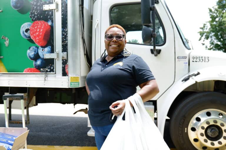 A smiling woman wearing sunglasses and a dark polo shirt stands in front of a truck with fruit images, holding white plastic bags. The truck door is open, and it’s a sunny day.