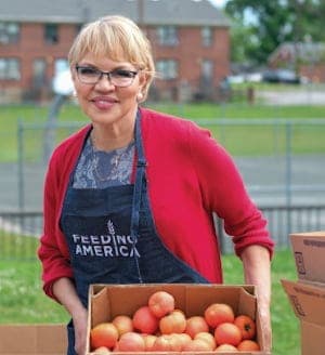 A woman wearing glasses and a Feeding America apron smiles while holding a box of tomatoes outdoors, with buildings and grass visible in the background.