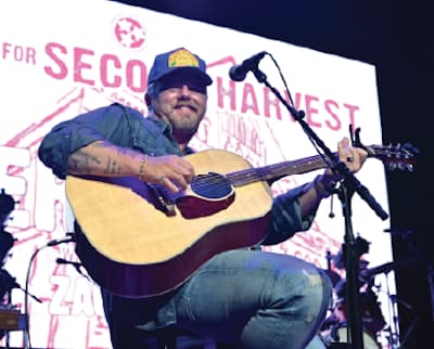 A man wearing a blue shirt and cap plays an acoustic guitar and sings into a microphone on stage. A large sign behind him reads SECOND HARVEST.