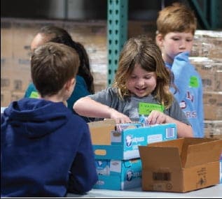Four children sort items into boxes at a table in what appears to be a warehouse or storage facility. One girl in front is smiling as she works, while the others focus on their tasks.