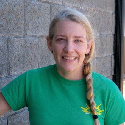 A woman with long blonde hair in a braid, wearing a green T-shirt, stands in front of a gray brick wall and smiles at the camera.