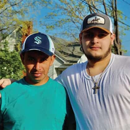 Two men stand closely side by side outdoors, one wearing a teal shirt and blue baseball cap, the other in a white shirt, chain necklace with a cross, and a trucker hat. Trees and a house appear in the background.