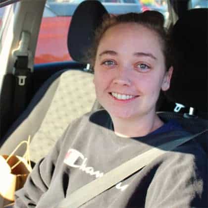 A young woman with light skin and brown hair pulled back smiles while sitting in the drivers seat of a car, wearing a black sweatshirt and a seatbelt. A brown bag is on the passenger seat.