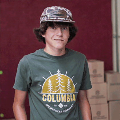 A young person with curly brown hair wears a camo-patterned cap and a green Columbia Sportswear t-shirt. They stand indoors in front of stacked cardboard boxes and a red wall, smiling slightly at the camera.